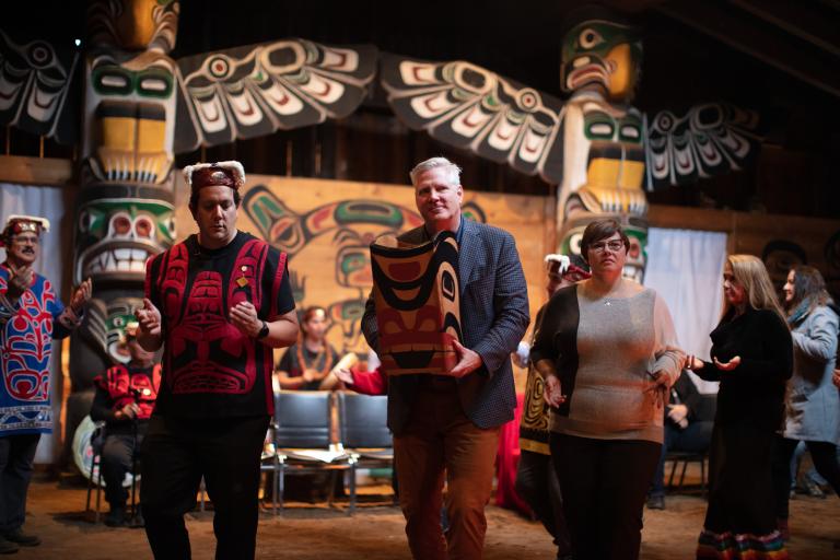 Two men and a woman dance side by side. One man is wearing a red vest and a headdress. The other man, in a suit jacket, carries a bentwood box carved in west coast Indigenous style. In the background are two larger totem poles and people dressed in regalia. Partially obscured.