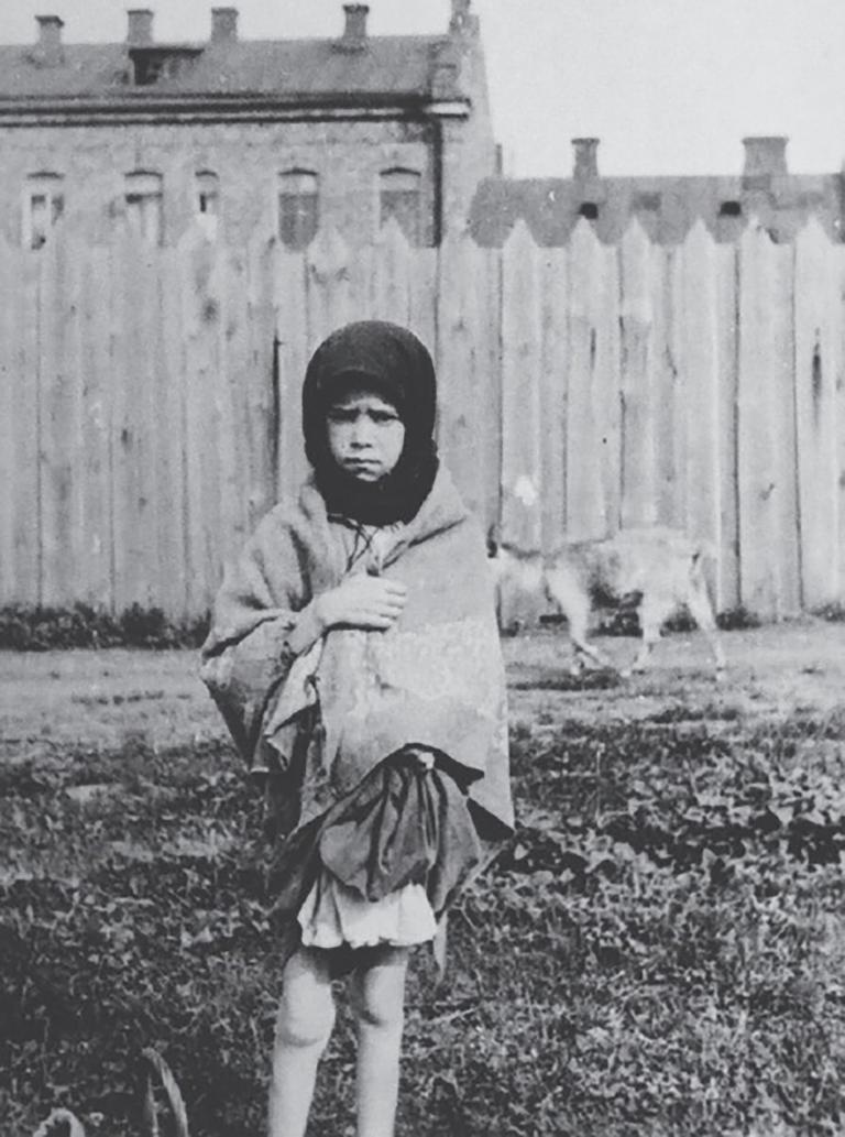 A young girl stands facing the camera with a sorrowful expression, clutching a shawl around her. Partially obscured.