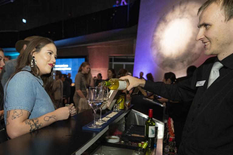 A woman waits for a drink at a bar in a museum space, while a bartender pours glasses of wine. Partially obscured.