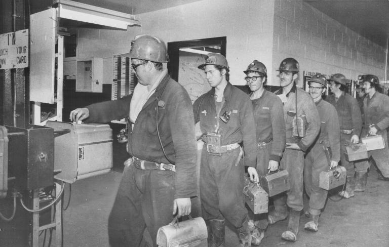 A line of miners hold lunch pails as they punch a time clock. Partially obscured.