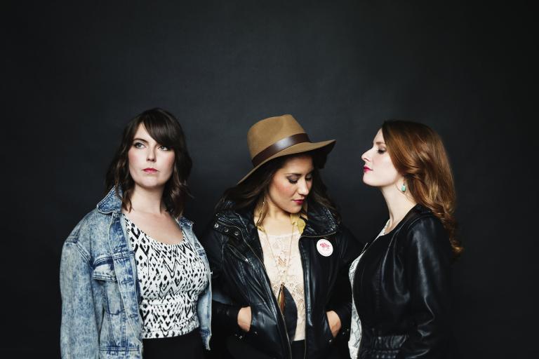 Three women pose in front of a black wall. Partially obscured.