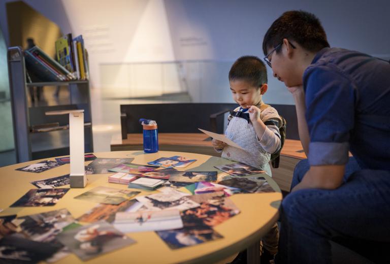 A young child interacting with photos on a table. An adult sits beside him. Partially obscured.