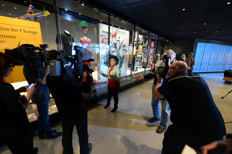 A young woman speaks while surrounded by camerapersons and photographers. Beside her is a large glass display case containing various objects, including a dress and tuxedo. 