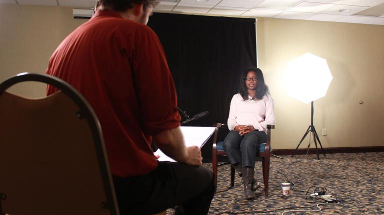 A man sits in a chair with his back facing the viewer. Sitting across from him in a chair is a young woman. A microphone is positioned in front of her. 