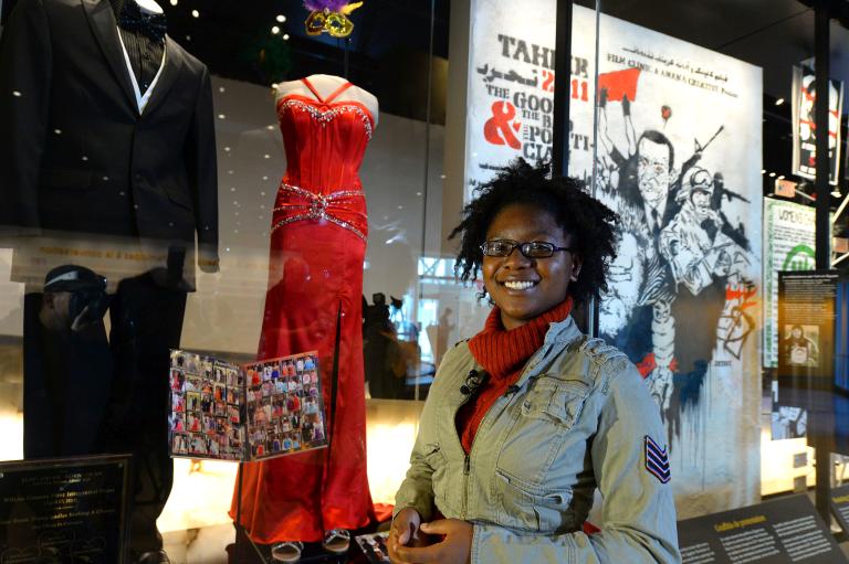 A smiling woman stands in front of a red prom dress and a black tuxedo mounted on mannequins. Both the dress and suit are on display behind a glass case. Partially obscured.