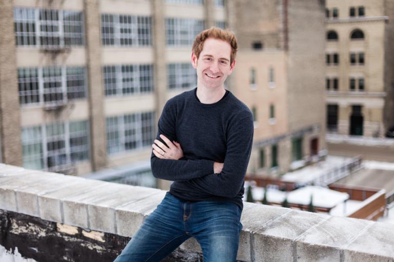 A man sits on the interior ledge of a rooftop. There are high-rise buildings in the background. Partially obscured.