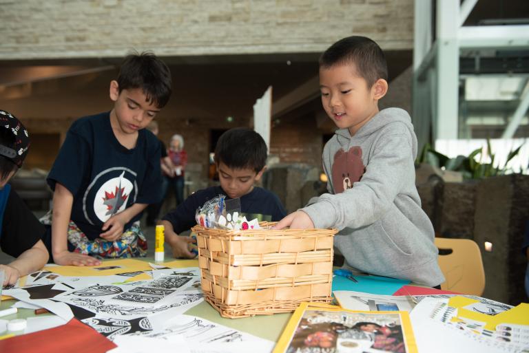 A child reaches into a basket of craft supplies while others work on their projects at a table. Partially obscured.