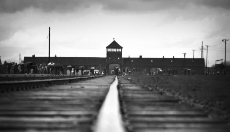 Train tracks lead to a distant building recognizable as the Auschwitz-Birkenau gate house. Partially obscured.