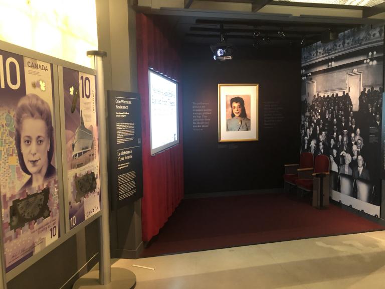 A museum exhibit that resembles an old-fashioned movie theatre. On one side are large representations of a $10 Canadian banknote featuring a woman’s face. Partially obscured.