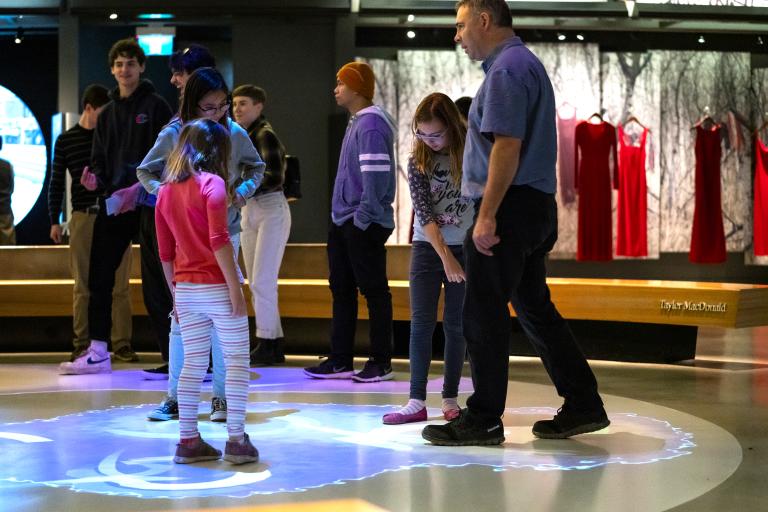 A crowd of people in a museum gallery. There are colourful light bubbles on the floor beneath them. Partially obscured.