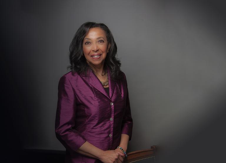 A portrait of a Black woman with long hair wearing a plum-coloured jacket. Partially obscured.