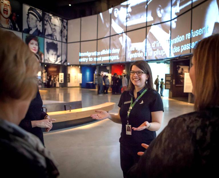 A female tour guide stands with outstretched arms in a museum gallery. Several tour participants are visible in the foreground. Partially obscured.