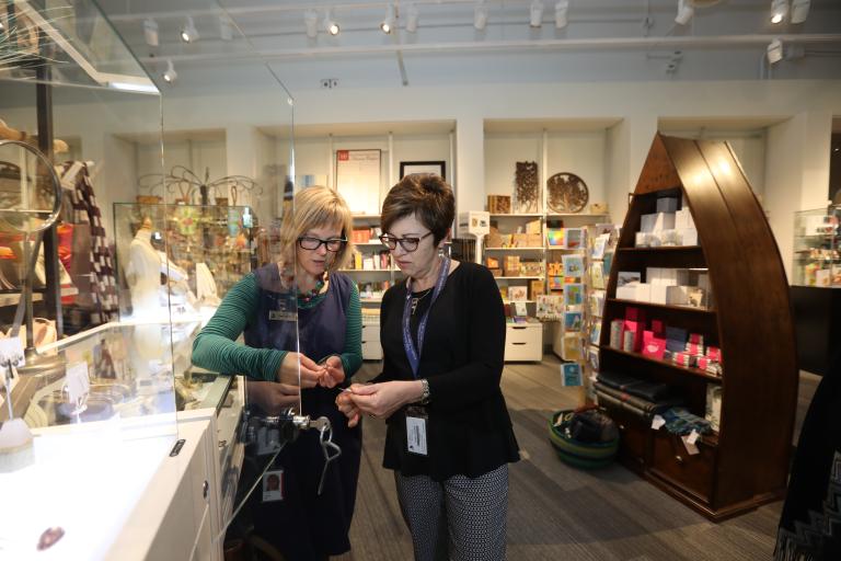 Two women look at a piece of jewellery. They are standing in a store full of colourful merchandise. Partially obscured.