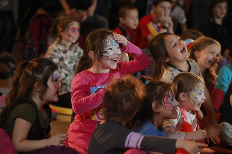 A group of children with playfully painted faces laughing and gesturing. Partially obscured.