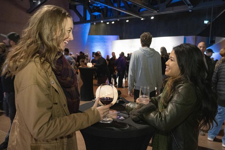 Two smiling women drink wine in a large room full of people. Partially obscured.