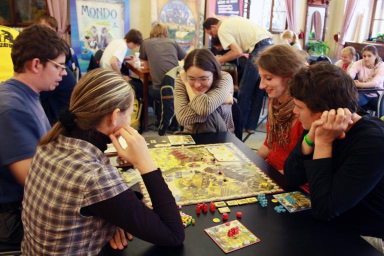 Three young women and two young men sit around a table playing a board game. There are other people playing board games at tables in the background. Partially obscured.