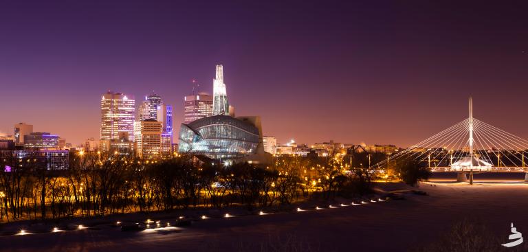 A city skyline at night. In the foreground is a river lined by trees, a glowing glass museum building and a bridge. Partially obscured.