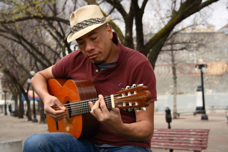 A man wearing a fedora plays a guitar while sitting outside on a bench. There are trees in the background. Partially obscured.