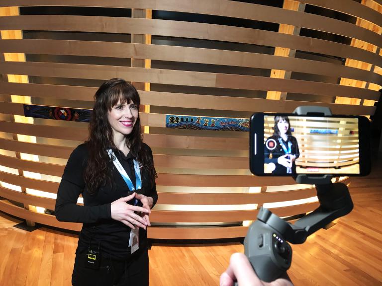 A smiling woman with light skin is filmed on a cell phone. She is wearing a lanyard with the words “Be inspired” and standing in front of an exhibit element made of horizontal strips of wood, some decorated with Indigenous art. Partially obscured.
