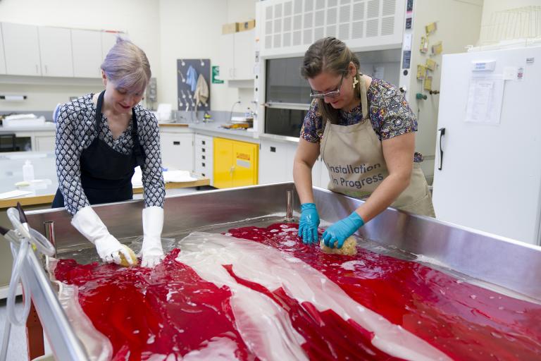 Two women wearing protective gloves and aprons wash a Canadian flag in a large sink.