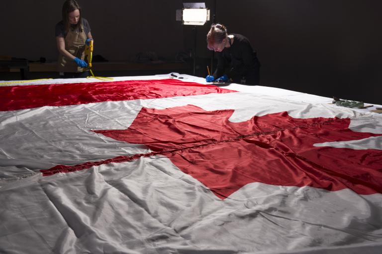 Two women wearing protective gloves and aprons working around a large Canadian flag, which lies flat on a table.