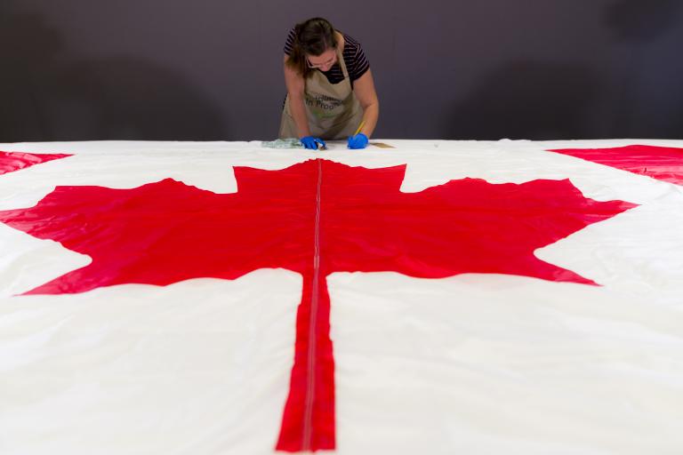 A woman wearing protective gloves and an apron takes notes behind a Canadian flag, which lies flat on a table in the foreground.