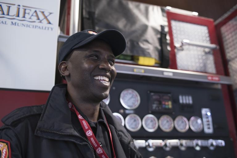 A smiling man stands in front of a fire truck. Partially obscured.