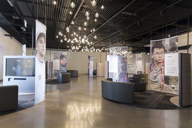 A museum display incorporating large portraits of diverse people, video screens and comfortable seating. 
