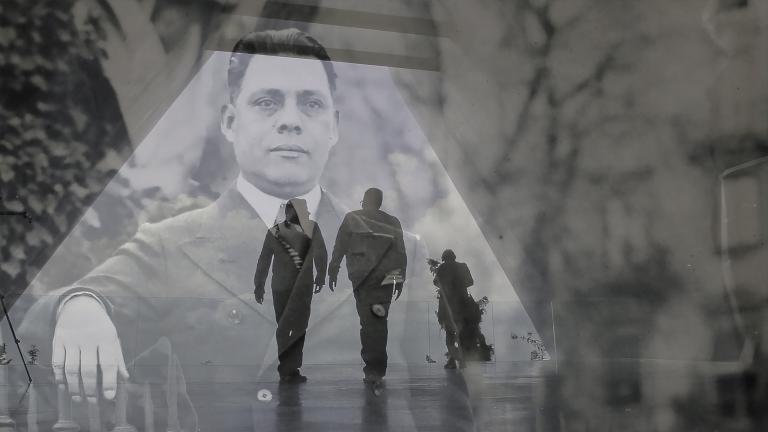 A translucent historical image of a man in a suit layered over a contemporary photo showing the dark outline of two men walking in a hallway. Partially obscured.