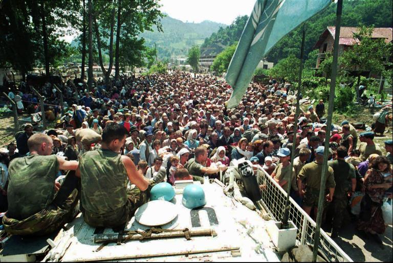 Two uniformed soldiers sit on a large vehicle, overlooking a densely packed crowd of people that stretches into the distance. Partially obscured.