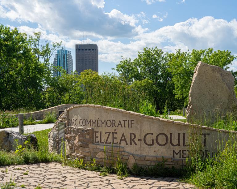 Rock and masonry sculpture shaped like a curved wall emblazoned with with large text reading “Parc commémoratif Elzéar Goulet.”