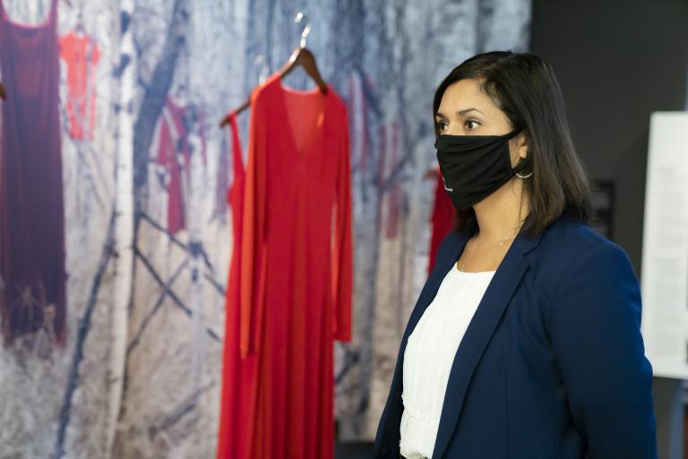 A woman wearing a black face mask stands in front of a museum exhibit consisting of red dresses hanging in front of a woodland background. Partially obscured.