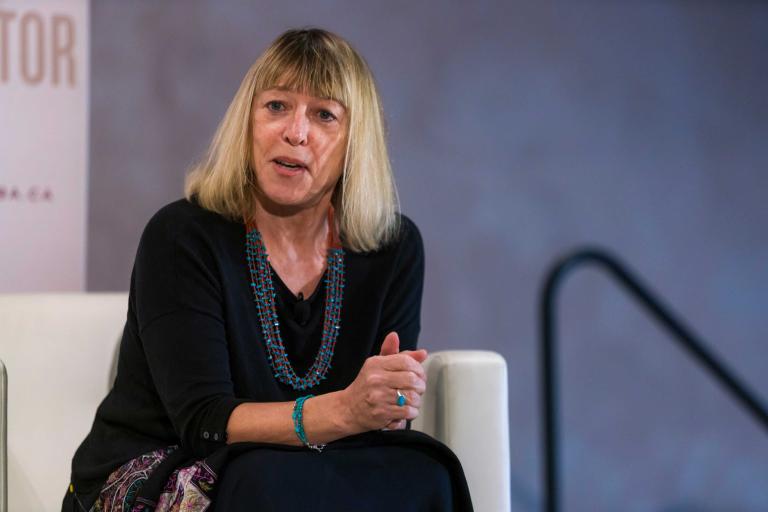 A woman sitting on a chair with hands clasped looks thoughtfully ahead, as if answering a question. Partially obscured.