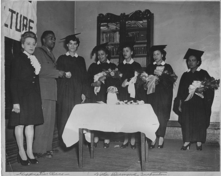 A black and white image of Viola Desmond standing next to a man in uniform. Five women wearing graduation caps and gowns and holding flowers stand to their left. One woman is accepting a scroll from the man.