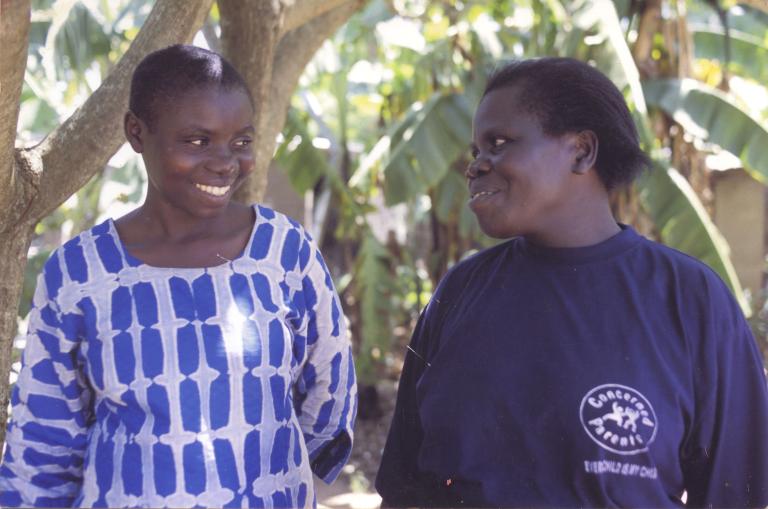 Two women are standing in front of tropical trees, looking at each other and smiling. The woman on the right is wearing a sweater that bears the logo of the Concerned Parents Association.