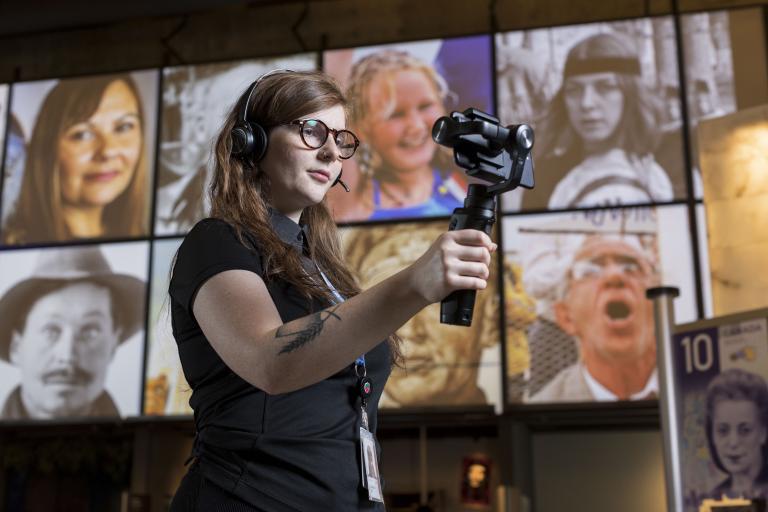 A woman wearing headphones, holding a mobile device on a handle, stands in a Museum gallery. Partially obscured.