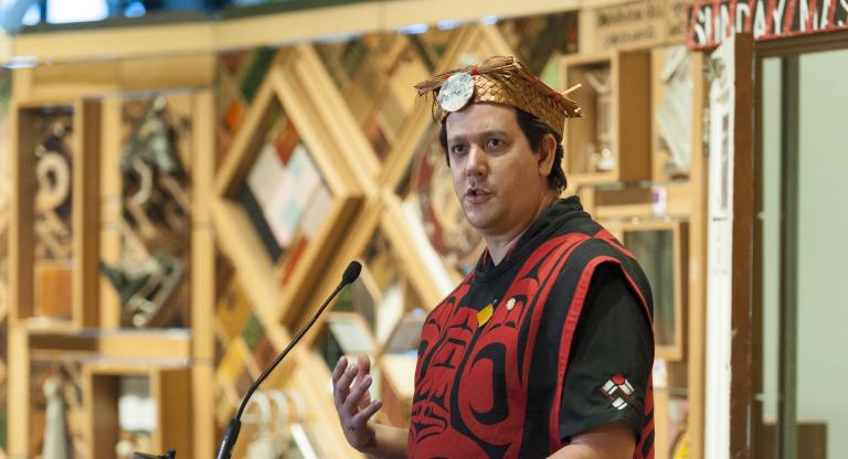 A man wearing traditional Indigenous regalia and headdress stands in front of an art installation made of wooden panels with embedded objects. Partially obscured.