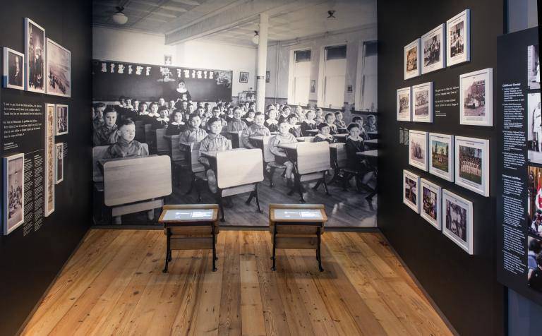 A museum exhibit that includes small school desks in front of a large photographic background of Indigenous children sitting at their desks in a classroom. Partially obscured.