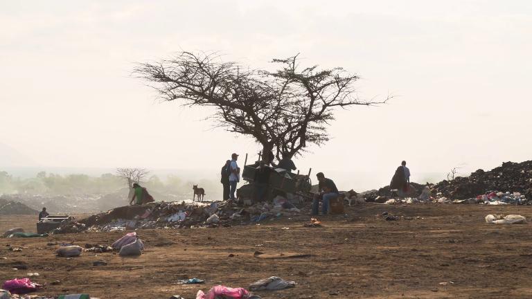 Several people and one dog sitting and standing in a landfill. A tree sits in the centre of the image and garbage is strewn on the ground. Partially obscured.