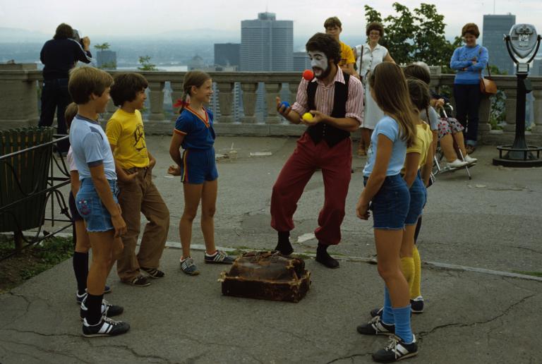 A man with a painted face juggles outdoors, as smiling children watch. A city skyline appears behind him at a distance.