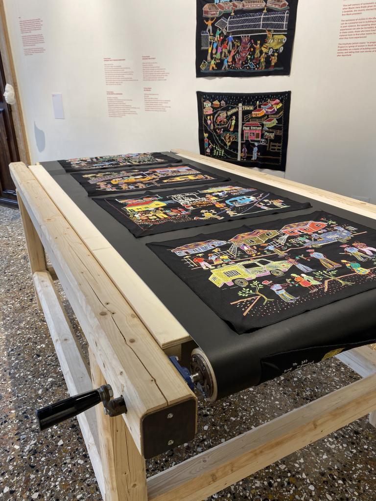 A series of colourful embroidery panels on black fabric displayed on a wooden hand-cranked conveyer belt table and on the wall of a museum.