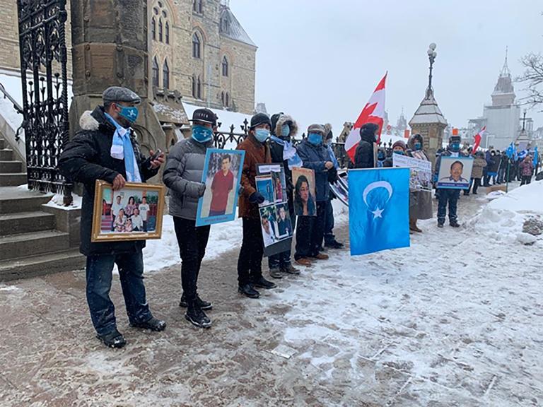  A line of people holding photographs and signs stand outside government buildings. Partially obscured.
