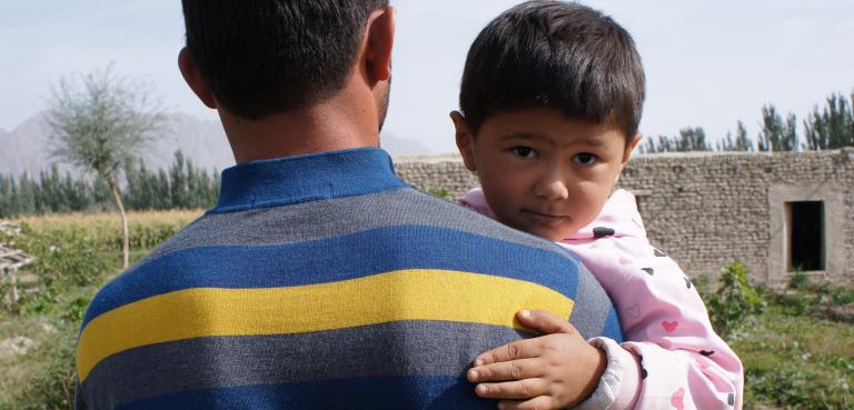 A young child being carried by an adult looks back over the adult’s shoulder. Partially obscured.