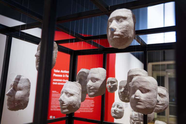 White casts of women’s faces hang by threads from the steel bars of a large cage. Partially obscured.