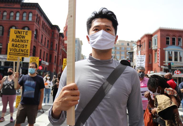 A man wearing a cloth mask and holding a sign, walking with a group of protestors carrying signs about ending racist violence and anti-Asian racism. Partially obscured.