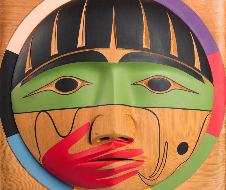 A carved wooden box, showing the carved face of a person with a painted red hand over their mouth. Partially obscured.