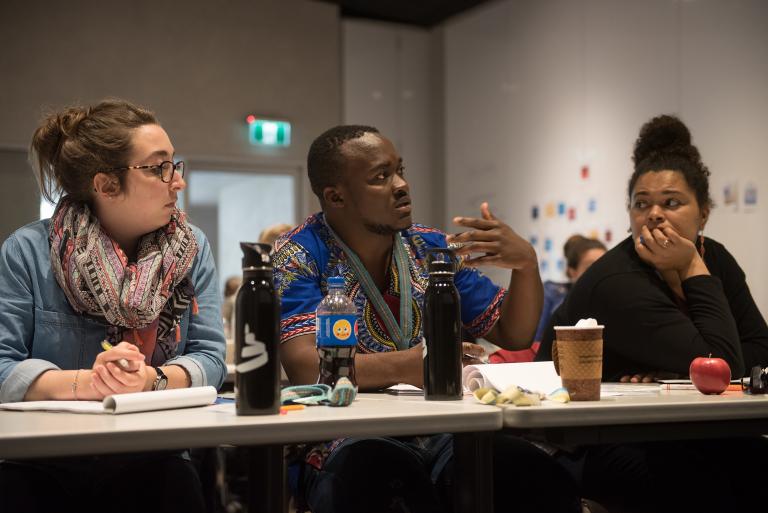 Three people sit at a table covered with water bottles, coffee cups and pads of paper. Partially obscured.