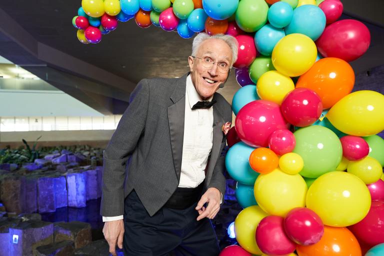 A performer wearing a tuxedo stands in front of a display of multi-coloured balloons. Partially obscured.