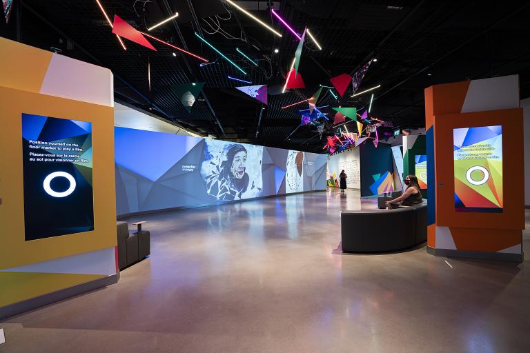 A museum gallery featuring artwork projected onto large screens and colourful geometric shapes. Partially obscured.