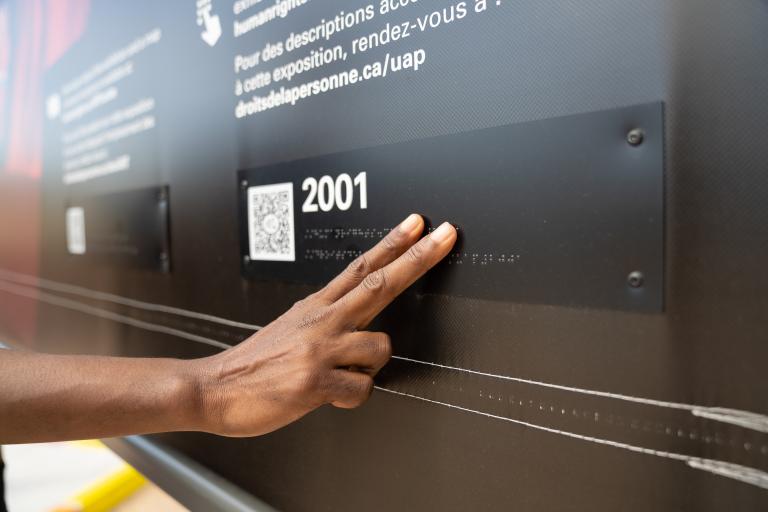 A hand touches braille letters on a museum exhibit. Partially obscured.